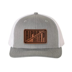 May include: A grey and white trucker hat featuring a rectangular brown leather patch on the front. The patch is embossed with the text ALPHA DELTA PI, the Greek letters ΑΔΠ, and the year 1851, separated by a diagonal line across the center.