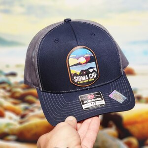 May include: Navy blue trucker hat with a gray mesh back and a patch featuring a mountain landscape with the text "SIGMA CHI". The hat has a curved brim and an adjustable strap.