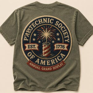 May include: Olive green t-shirt featuring a circular patch with the text "PRRUTECHNIC SOCIETY OF AMERICA" and "ANNUAL GRAND DISPLAY". The patch includes a firework design, stars, and the year "1776".