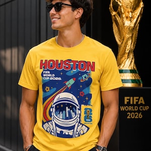 May include: A yellow t-shirt with a graphic promoting the FIFA World Cup 2026 in Houston. The design features an astronaut, a rocket, and the text "HOUSTON FIFA WORLD CUP 2026". A gold trophy is in the background.