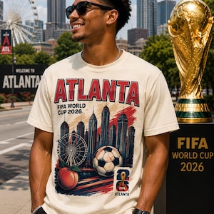 May include: Cream-colored t-shirt with the word "ATLANTA" in red, featuring a soccer ball, Ferris wheel, and peach graphic. The shirt promotes the FIFA World Cup 2026. A golden trophy is in the background.
