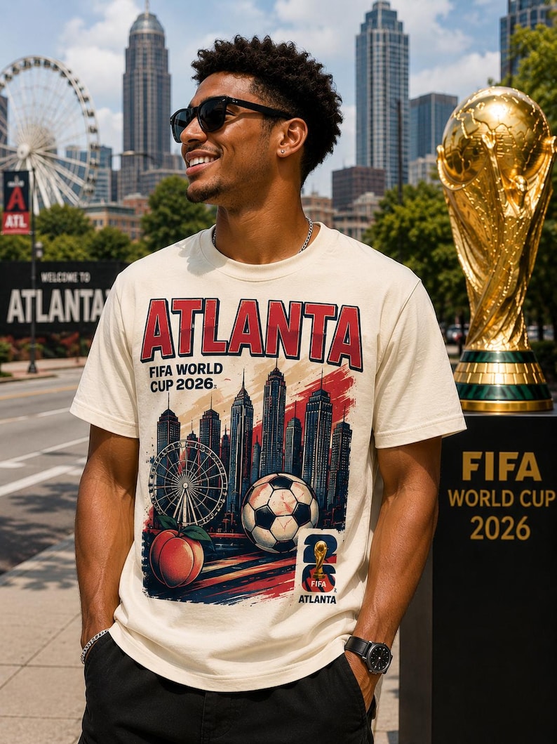 May include: Cream-colored t-shirt with the word "ATLANTA" in red, featuring a soccer ball, Ferris wheel, and peach graphic. The shirt promotes the FIFA World Cup 2026. A golden trophy is in the background.