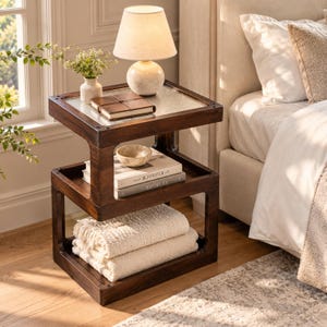 May include: A dark wood three-tier side table with a glass top, featuring a lamp, a small vase with flowers, a notebook, a bowl, two books titled The Kinfolk and At Home, and two folded cream-colored blankets on the bottom shelf.