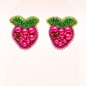 May include: A pair of pink and green beaded earrings shaped like raspberries. The earrings are displayed on a wooden stand.