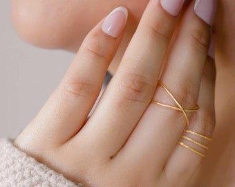 Puede incluir: Primer plano de una mano con tres anillos de oro. Los anillos están apilados en el dedo anular. El anillo superior es una banda cruzada delicada. Los dos anillos inferiores son finos y texturizados.