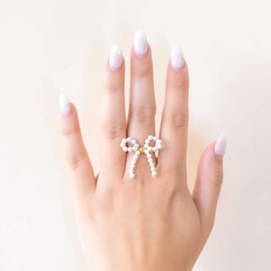 May include: A hand wearing a ring with a pearl bow design. The hand is also wearing a bracelet with gold and white beads.