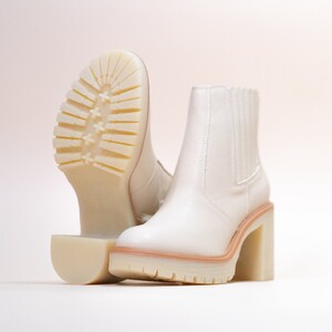 May include: A pair of off-white ankle boots featuring a smooth upper, elastic side panels, and a chunky block heel. The thick, lugged rubber sole provides traction, with one boot positioned to display the textured tread pattern on the bottom.