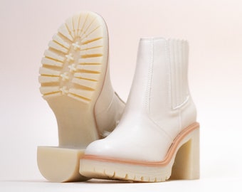 Puede incluir: Un par de botines color blanco hueso con parte superior lisa, paneles laterales elásticos y un tacón de bloque grueso. La suela de goma gruesa y dentada proporciona tracción, con una bota colocada para mostrar el patrón de la suela.