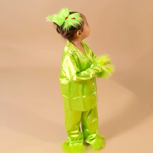 May include: A child's lime green pajama set with white trim, featuring a button-down top and pants. The outfit includes matching fluffy trim on the cuffs, ankles, and a hair accessory. The ensemble is a festive costume.