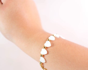 May include: A gold chain bracelet with white heart-shaped beads. The bracelet is on a person's wrist.