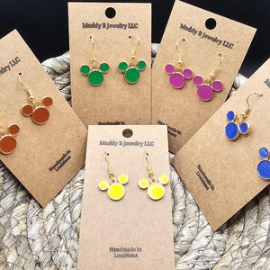 May include: A selection of colourful earrings, including orange, green, pink, blue, and yellow, presented on brown cards. Each pair features a three-circle design with gold-coloured hooks. The cards are labelled "Maddy B Jewelry LLC" and "Handmade in Louisiana."