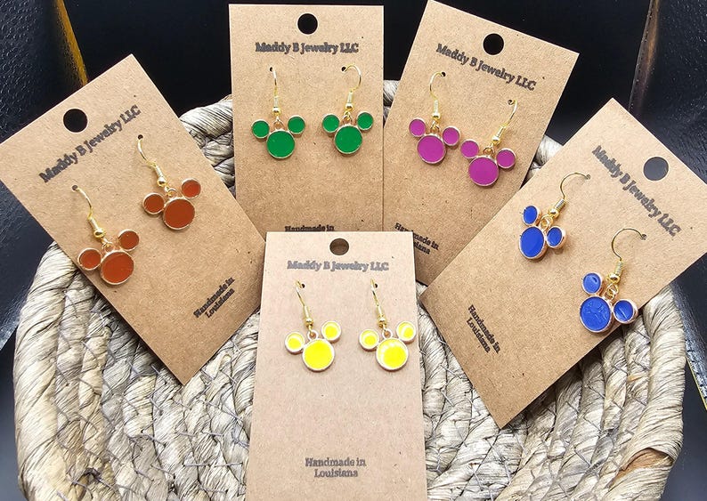 May include: A selection of colourful earrings, including orange, green, pink, blue, and yellow, presented on brown cards. Each pair features a three-circle design with gold-coloured hooks. The cards are labelled "Maddy B Jewelry LLC" and "Handmade in Louisiana."