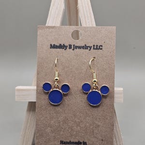 May include: Gold-coloured drop earrings with a three-circle design in royal blue enamel. The earrings are displayed on a brown card with the text "Maddly B Jewelry LLC" and "Handmade in Louisiana".