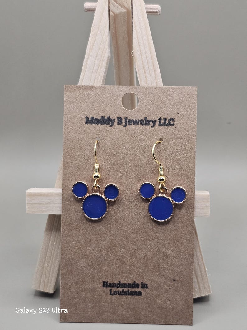 May include: Gold-coloured drop earrings with a three-circle design in royal blue enamel. The earrings are displayed on a brown card with the text "Maddly B Jewelry LLC" and "Handmade in Louisiana".