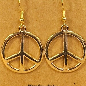 May include: A pair of gold-colored peace sign earrings. The earrings are round with a peace symbol in the center. The earrings are hanging from gold-colored hooks. The text "Maddy B Jewelry LLC" is at the top and "Handmade in Louisiana" is at the bottom.