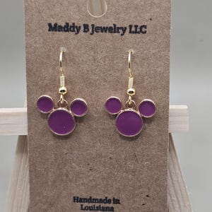 May include: Gold-coloured drop earrings featuring a purple circular design. The earrings are presented on a brown card with the text "Maddy B Jewelry LLC" and "Handmade in Louisiana".