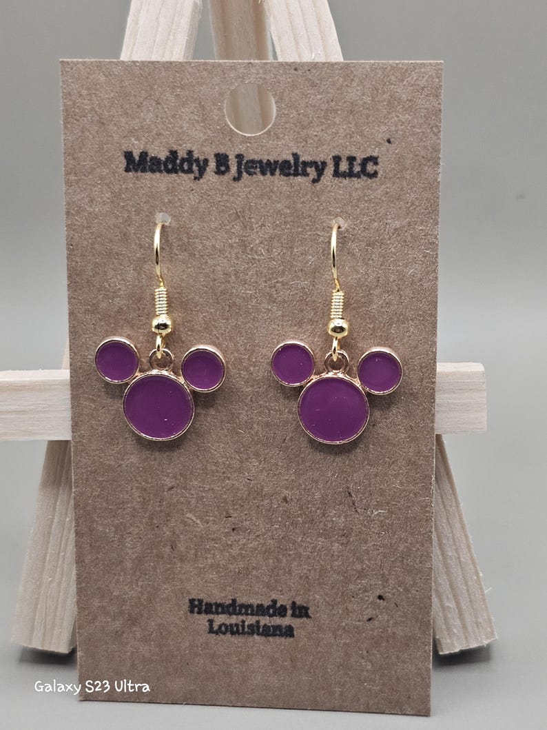 May include: Gold-coloured drop earrings featuring a purple circular design. The earrings are presented on a brown card with the text "Maddy B Jewelry LLC" and "Handmade in Louisiana".