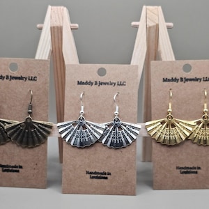 May include: Three pairs of fan-shaped earrings in bronze, silver, and gold tones are displayed on brown cards. Each card is labeled "Maddy B Jewelry LLC" and "Handmade in Louisiana."