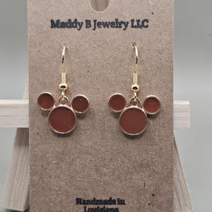 May include: Gold-coloured dangle earrings featuring a three-circle design in a reddish-brown shade. The earrings are presented on a brown card with the text "Maddy B Jewelry LLC" and "Handmade in Louisiana".