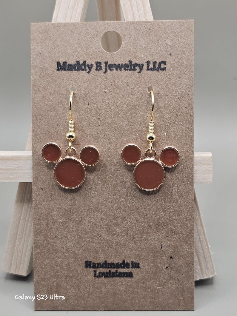May include: Gold-coloured dangle earrings featuring a three-circle design in a reddish-brown shade. The earrings are presented on a brown card with the text "Maddy B Jewelry LLC" and "Handmade in Louisiana".