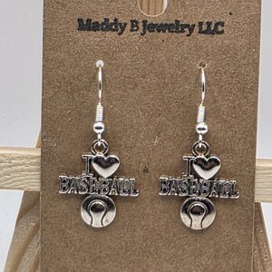 May include: A pair of silver-toned dangle earrings featuring a charm with a heart, the text I BASEBALL, and a baseball icon. The earrings are attached to a brown display card printed with Maddy B Jewelry LLC and Handmade in Louisiana.