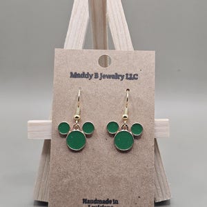 May include: Gold-coloured drop earrings featuring a green enamel Mickey Mouse design. The earrings are presented on a brown card with the text "Maddy B Jewelry LLC" and "Handmade in Louisiana".