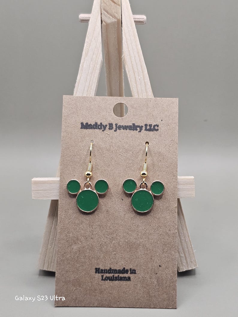 May include: Gold-coloured drop earrings featuring a green enamel Mickey Mouse design. The earrings are presented on a brown card with the text "Maddy B Jewelry LLC" and "Handmade in Louisiana".