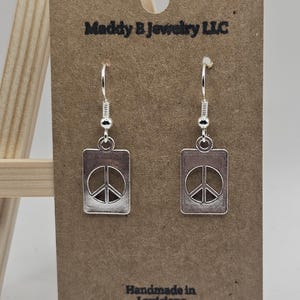 May include: Silver-toned peace sign earrings with a rectangular pendant. The earrings are displayed on a brown card with the text "Maddy B Jewelry LLC" and "Handmade in Louisiana".