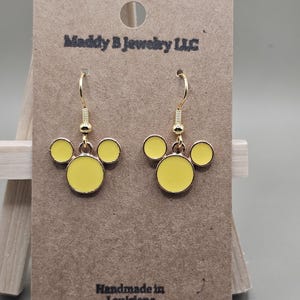 May include: Gold-coloured drop earrings featuring a yellow Mickey Mouse-style design. The earrings are presented on a brown card with the text "Maddy B Jewelry LLC" and "Handmade in Louisiana".