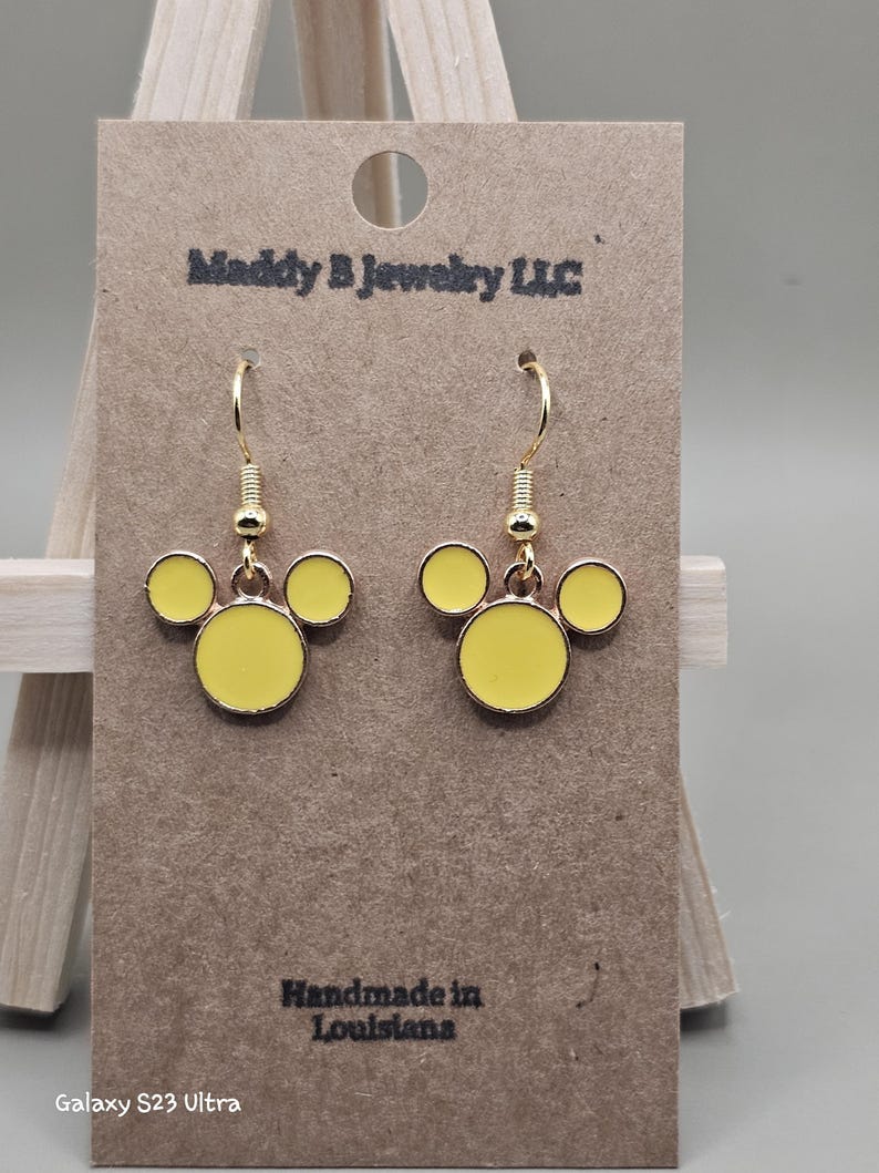 May include: Gold-coloured drop earrings featuring a yellow Mickey Mouse-style design. The earrings are presented on a brown card with the text "Maddy B Jewelry LLC" and "Handmade in Louisiana".