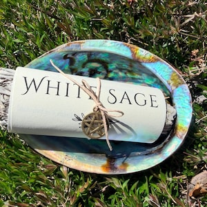 Puede incluir: Un paquete de salvia blanca envuelto en papel blanco con la inscripción "WHITE SAGE", dentro de una concha de abulón iridiscente. Un dije dorado con un pentagrama cuelga del hilo. La concha descansa sobre hierba verde.