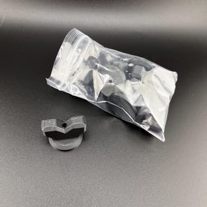 以下が含まれることがあります： いくつかの小さな黒いプラスチック部品が入った透明なビニール袋。1つの部品は分離されており、上部が湾曲し、円形のベースを持つ濃い灰色の部分です。アイテムは濃い灰色の表面にあります。