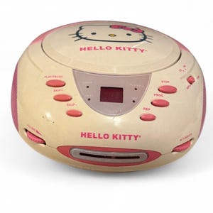 Puede incluir: Un reproductor de CD y radio portátil Hello Kitty de forma redonda, color crema con detalles en rosa. El dispositivo cuenta con un compartimento de carga superior para CD con un gráfico de Hello Kitty, pantalla digital, botones rosas y diales laterales.