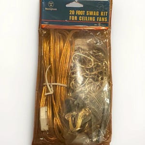 Könnte beinhalten: Ein Westinghouse 20-Fuß-Aufhängungsset für Deckenventilatoren in der Original-Kunststoffverpackung. Das Set enthält ein langes goldfarbenes Stromkabel mit weißem Stecker sowie eine goldfarbene Metallkette mit Zubehör für die Montage.