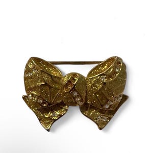 May include: A gold-toned bow brooch featuring a textured, glittery surface finish. The bow is embellished with small, clear, round inset stones arranged in diagonal lines across the loops and the central knot, with a horizontal pin on the back.