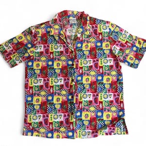 Op de afbeelding: Een overhemd met korte mouwen en een kleurrijk fruitpatroon. Het shirt heeft een collage van vierkanten met illustraties van citroenen, watermeloenen en ander fruit in rood, geel, blauw en groen. Een klein label is zichtbaar op de kraag.