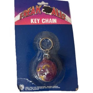 May include: A vintage Freak Alls key chain on its original packaging. The key chain features a purple, round charm with a yellow face. The packaging is blue and red with the text "FREAK ALLS KEY CHAIN".