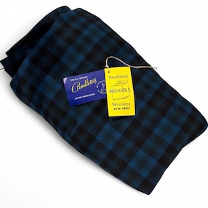 Może przedstawiać: Złożony wełniany koc w ciemnoniebiesko-czarną kratę. Dołączone są dwie metki: niebieska z napisem "This is a genuine Pendleton Always Virgin Wool" oraz żółta z napisem "Pendleton Washable Woolens Moth-Proof".