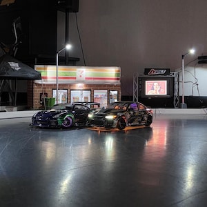 May include: Two miniature remote-controlled cars on a track, one dark blue and the other black with colourful accents. A miniature 7-Eleven store and a TV screen are in the background, with bright lighting.