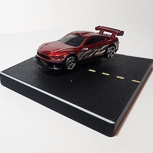 May include: A red toy car with a black and white racing stripe design on a black display base with a white border. The car has a rear spoiler and is on a black surface with yellow lines.