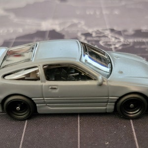 May include: A gray die-cast model car with black wheels. The miniature vehicle has a detailed interior, a rear spoiler, and a sunroof. The car's headlights are yellow, and the taillights are red. The model is a replica of a sports car.