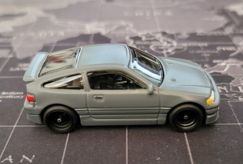 May include: A gray die-cast model car with black wheels. The miniature vehicle has a detailed interior, a rear spoiler, and a sunroof. The car's headlights are yellow, and the taillights are red. The model is a replica of a sports car.