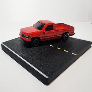 May include: A red toy pickup truck with a black interior and black wheels is displayed on a black surface with white lines.