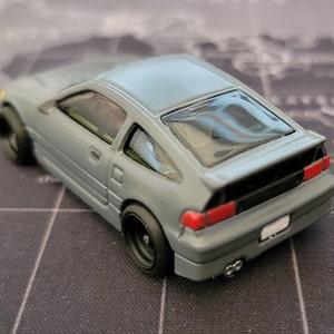 May include: A gray diecast model car with black wheels and a rear spoiler. The car has red taillights and a white license plate. The model is a coupe with a hatchback design.