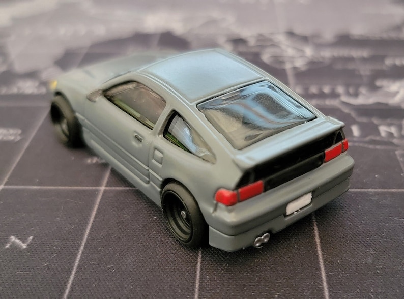 May include: A gray diecast model car with black wheels and a rear spoiler. The car has red taillights and a white license plate. The model is a coupe with a hatchback design.