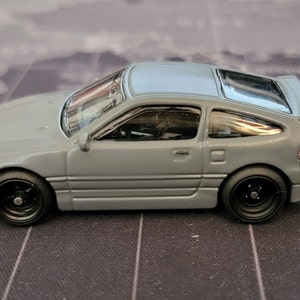 May include: A gray die-cast model car with a light blue roof and black wheels. The miniature vehicle has a spoiler and a detailed interior. The car is a coupe style and sits on a black surface.