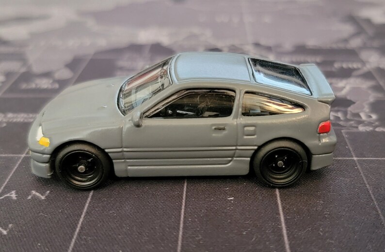 May include: A gray die-cast model car with a light blue roof and black wheels. The miniature vehicle has a spoiler and a detailed interior. The car is a coupe style and sits on a black surface.