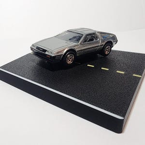 May include: A silver die-cast DeLorean model car with black and gold wheels, displayed on a black textured platform with yellow lane markings. The car is a detailed collectible item.