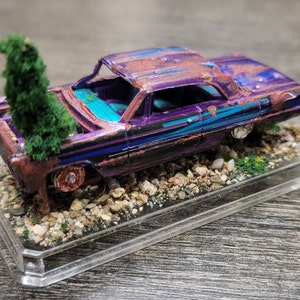 Può includere: Un modello in miniatura di auto viola e nera usurata, con accenti ruggine e turchese. Un piccolo albero verde spunta dal tetto aperto dell'auto. Il modello poggia su una base trasparente in acrilico con ghiaia e piante in miniatura.