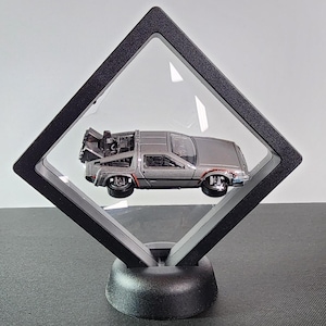 May include: A silver DeLorean time machine toy car displayed in a black diamond-shaped display case with a clear acrylic front. The case has a black base.